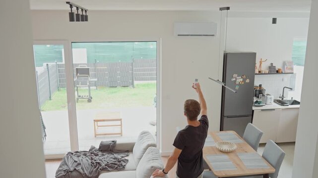 Man using remote control to adjust air conditioner in bright modern living room. Person adjusting temperature for cooling during hot summer days. Concept of home comfort