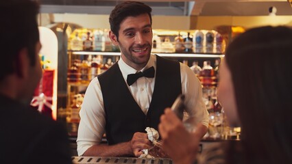 Focusing smiling smart bartender serving gin fizz or tonic soda at luxurious bar, engaging lively conversation with VIP customer couple at back view in atmosphere at lighting warm nightclub. Vinosity.