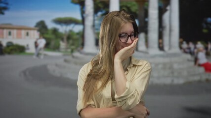 Young blonde woman wearing beige shirt adjusting glasses and touching cheek on street; embarrassment.