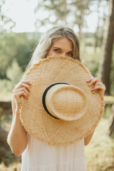 Young blonde woman hiding her face behind a large straw hat in nature