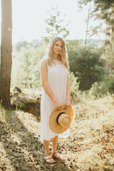 Blonde woman holding straw hat enjoying sunny forest landscape