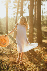 Woman walking in forest holding straw hat and bag at sunset