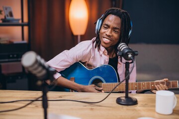 African American man musician recording podcast, wearing headphones, guitar in hand, surrounded by...