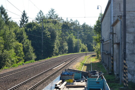 A panoramic view of two parallel railway tracks curving away into the distance, with a lush forest on one side and a concrete industrial building on the other, under a clear sky