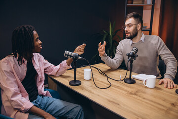Two male podcasters, one african american and one caucasian, talking and recording a podcast in a...