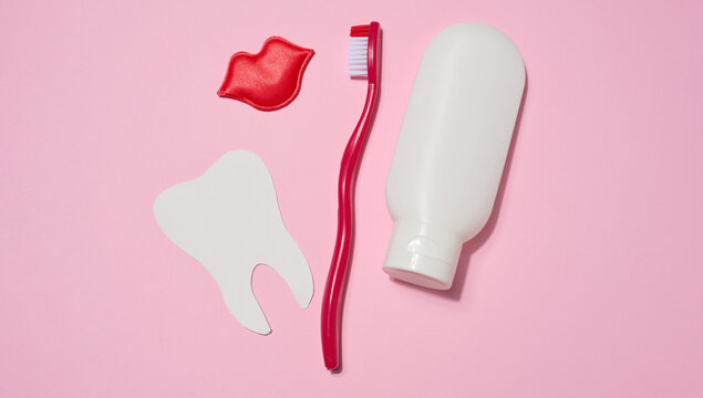 Red toothbrush, red tube of toothpaste, red decorative lips and white paper tooth cut out on pink background.