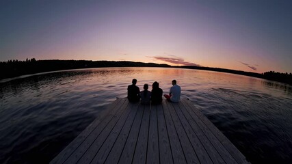 A wide-angle video shot captures four people sitting on a dock, silhouetted against a serene sunset over a calm lake, evoking tranquility.