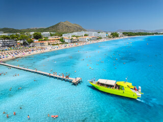 Aerial view of Alcudia beach featuring tourists swimming, sunbathing, and walking on the pier, with hotels and a tour boat. Mallorca, Balearic Islands, Spain. Top view of yacht, blue sea, sandy beach