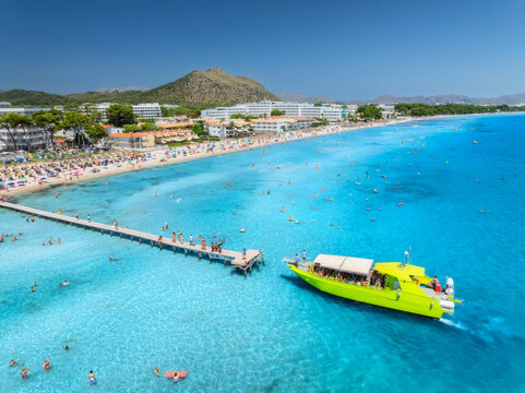 Aerial view of Alcudia beach featuring tourists swimming, sunbathing, and walking on the pier, with hotels and a tour boat. Mallorca, Balearic Islands, Spain. Top view of yacht, blue sea, sandy beach - Powered by Adobe