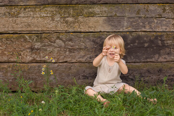 a happy handsome blond boy with long hair is sitting on the grass holding a daisy in his hands against the background of an old wooden wall