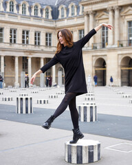 Happy stylish woman in black dress balancing on striped column at Palais Royal in Paris, France,...
