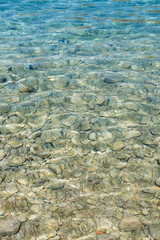 Clear turquoise water surface reveals smooth stones beneath, creating a tranquil underwater scene with reflections and gentle ripples, perfect for relaxation. Vertical background with copy space
