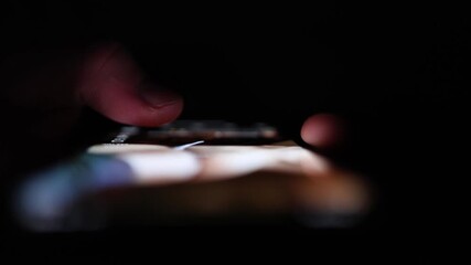 Glowing smartphone screen reflects on fingertips in the dark with blurred content evoking late-night scrolling digital overload and the quiet pull of technology before sleep