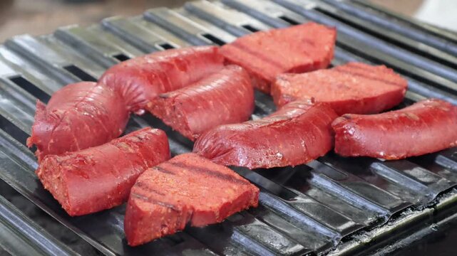 Traditional Turkish sausage (sucuk) being grilled on a charcoal barbecue. A person uses metal tongs to flip the sizzling sausages. Smoke rises as the meat cooks over open fire, creati