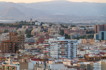Aerial view of Alicante city at dusk, showcasing a blend of modern and traditional architecture with distant mountains and a hazy skyline, enhancing the urban landscapes appeal.