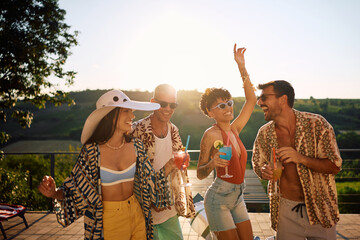 Multiracial group of happy friends drinking cocktails and dancing during summer party on patio.