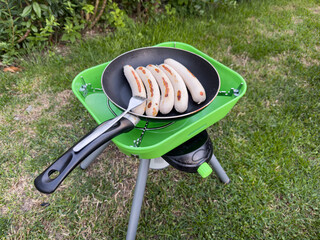 Cooking sausages in a frying pan on a portable camping stove, surrounded by green grass and outdoor scenery, showcasing outdoor culinary experience