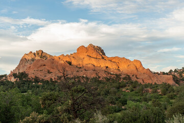 Fototapeta premium Warm golden-hour light illuminates red sandstone ridges above a forested hillside, set against a partly cloudy sky, showcasing dramatic geology and lush green foothill contrast.