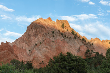 Sunlit red sandstone cliffs glow at golden hour above pine-covered foothills under a blue sky with wispy clouds, showcasing warm light on rugged geological textures.