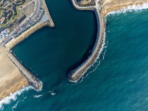 Aerial view of the entrance pier to Nazaré Marina, Portugal, highlighting the coastal pier structure and vibrant blue ocean waters under clear skies.