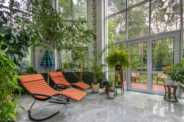 A cozy indoor garden with sun loungers, lush plants and large windows letting in sunlight.