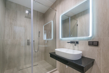 A modern bathroom with a glass shower, a stylish sink on a black countertop and a backlit mirror....