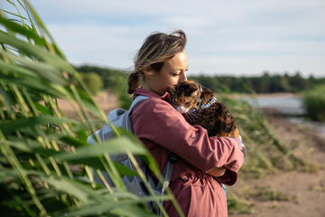 Woman kissing cuddling tense fluffed cat on walk by coast through tall grass. Pet owner caressing...