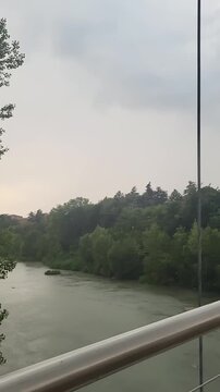 View of a suspension bridge with wet wooden planks and green trees under an overcast sky in the day. Budrio, Italy