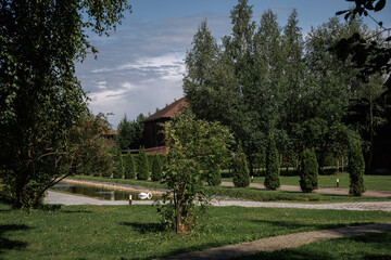 Tranquil park setting featuring lush greenery, a pathway, and a pond. Tall trees and manicured bushes create a serene environment perfect for relaxation and leisure activities.
