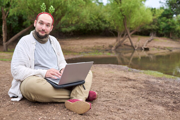 A young Hispanic man wearing a Christmas reindeer antler headband using a laptop while sitting on...