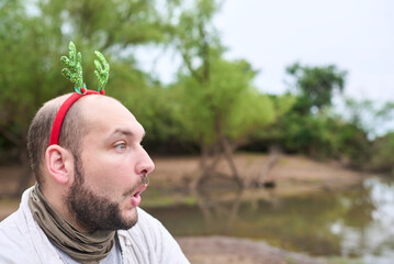 Portrait of a young man wearing a funny headband with Christmas reindeer antlers, outdoors in...