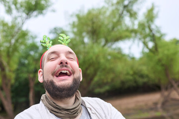 A young Hispanic man wearing a funny Christmas reindeer antler headband smiles with a funny expression outdoors, in a natural setting.