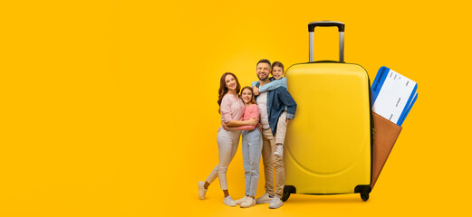 A cheerful family of four stands next to a bright yellow suitcase, happily engaging in travel planning. They are excited about their upcoming summer vacation and tourist activities.