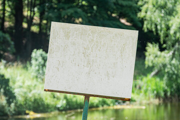 Large blank sign in an ancient Central European village setting, with a reflective body of water nearby, under natural and diffuse lighting The predominant colors are green, brown, and white