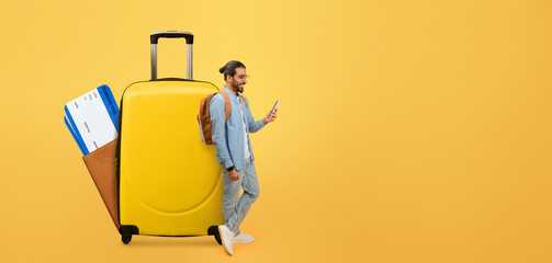 A traveler stands next to a bright yellow suitcase while using a smartphone. With travel tickets...