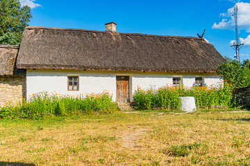 Quaint white cottage, lush greenery, dark brown shingled roof, multiple chimneys, gray wooden door,...
