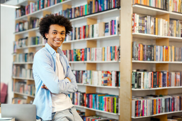 Intelligent African American student guy standing in library in front of bookshelves, happily...