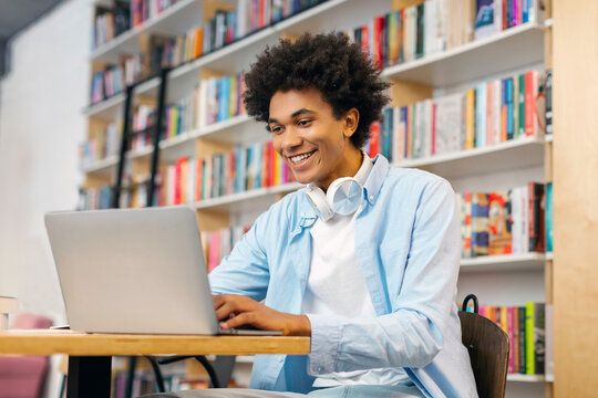 Happy Black male university student preparing for lecture, using laptop while studying in college library, using modern wireless tech and internet resources