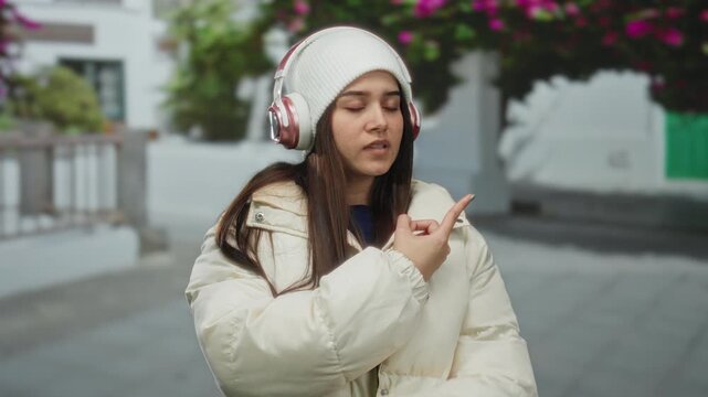 Young woman wearing headphones and a winter jacket listens to music on a peaceful street lined with flowering trees, enjoying a moment outdoors.