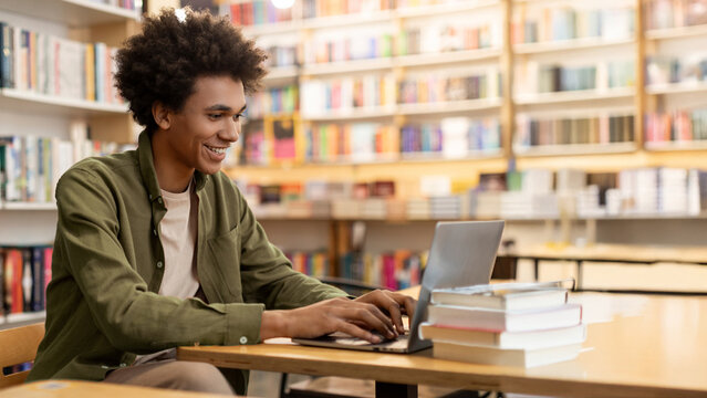 Smart Latin student guy sitting at desk in campus library using laptop and doing homework or preparing for exam