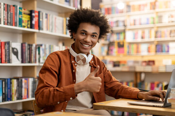 Happy black learner guy sitting at table in college library and showing thumbs up, enjoying study...