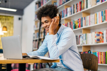 Cheerful black student guy studying in university library, using laptop with headphones and...