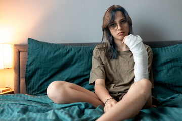 Teenage girl with cast on broken arm sitting on bed at home, recovering and using smartphone at...