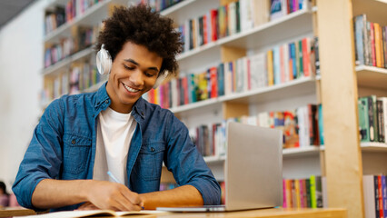 Positive African American student guy with headphones taking notes, having online lecture while...