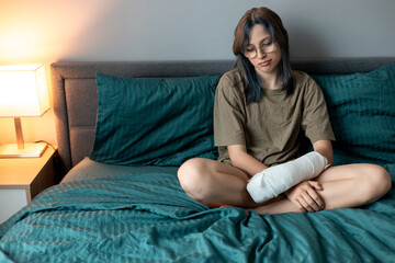 Teenage girl with cast on broken arm sitting on bed at home, recovering and using smartphone at...