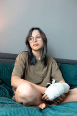 Teenage girl with cast on broken arm sitting on bed at home, recovering and using smartphone at...