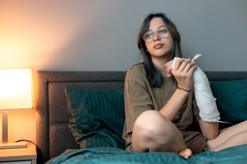 Teenage girl with cast on broken arm sitting on bed at home, recovering and using smartphone at...