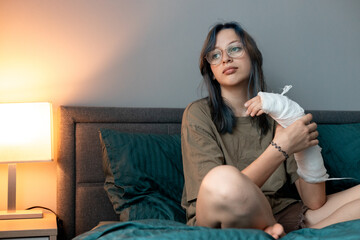 Teenage girl with cast on broken arm sitting on bed at home, recovering and using smartphone at...