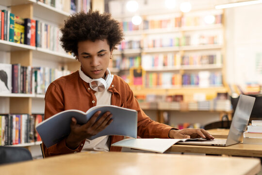 Concentrated black guy engrossed in study, reading book and using laptop, sitting in university library, preparing for lecture or tests