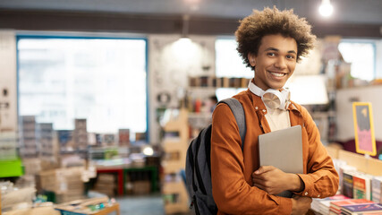 Excited African American guy wearing backpack and holding laptop, standing in college library at...
